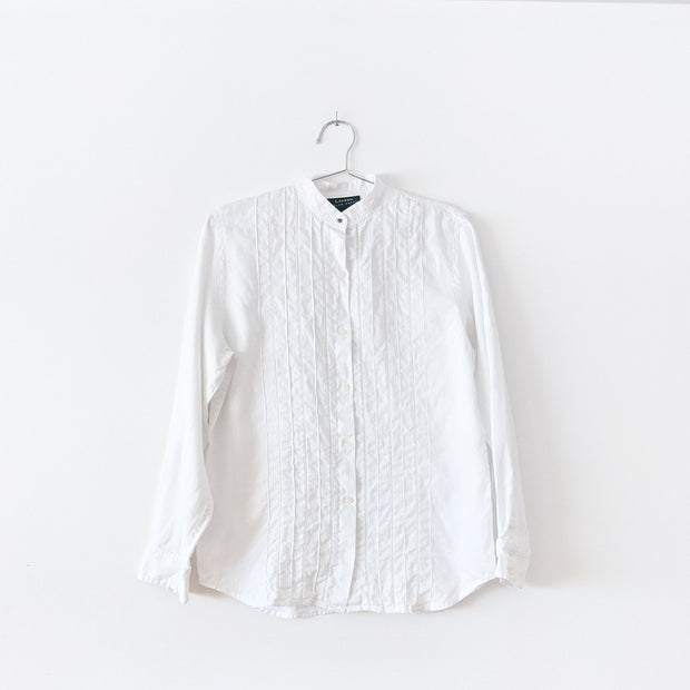 White linen long sleeve button-up shirt with mandarin collar and brass button. There is vertical pleating and white ladder lace at the center front. Secondhand Lauren Ralph Lauren, from Fold and Fray.