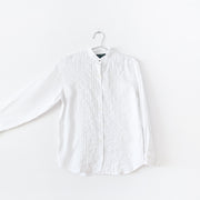 White linen long sleeve button-up shirt with mandarin collar and brass button. There is vertical pleating and white ladder lace at the center front. Arm extended to show sleeve. Secondhand Lauren Ralph Lauren from Fold and Fray.