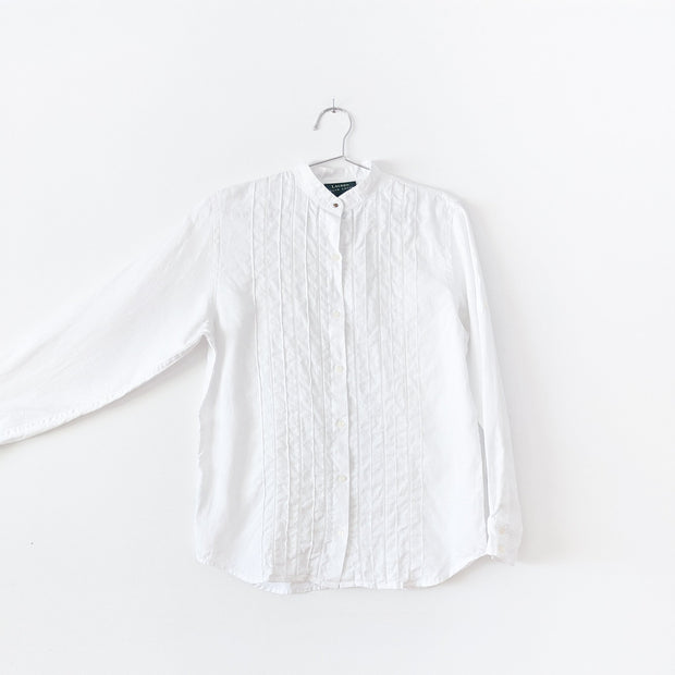 White linen long sleeve button-up shirt with mandarin collar and brass button. There is vertical pleating and white ladder lace at the center front. Arm extended to show sleeve. Secondhand Lauren Ralph Lauren from Fold and Fray.