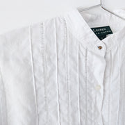 Close-up of mandarin collar with a spherical brass button, and white ladder lace trim and vertical pleating on a white linen long sleeve button-up shirt. Secondhand Lauren Ralph Lauren from Fold and Fray.