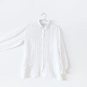 White linen long sleeve button-up shirt with vertical pleats, six inch side slits, and white ladder lace trim down the center on the front. Arm extended to show sleeve.  Liz Claiborne Lizsport. Secondhand from Fold and Fray.