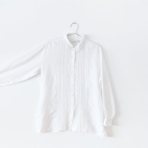 White linen long sleeve button-up shirt with vertical pleats, six inch side slits, and white ladder lace trim down the center on the front. Arm extended to show sleeve.  Liz Claiborne Lizsport. Secondhand from Fold and Fray.