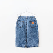 Back of 1980’s style high-waisted blue denim acid wash pencil skirt, brown patch on back pocket, back slit at bottom.