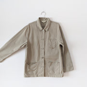 Cotton, field-style, barn jacket, chore coat in Sand Beige (like a greige, tan). Straight fit coat with three front patch pockets, metal buttons, a collar and detailed tapered sleeve cuffs. Arm extended to show sleeve width. Eddie Bauer Women's. Secondhand, from Fold and Fray.