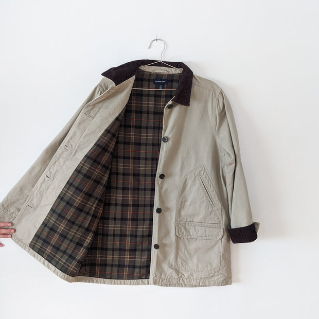 Cotton canvas field-style barn jacket, chore coat in Sand Beige (like a greige tan) opened to show the inside. It has dark brown buttons and dark brown corduroy lined collar and cuffs. The inside of the jacket's torso is lined with green plaid flannel. This jacket has four exterior pockets: two lower patch pockets with flaps and two upper angled welt pockets. Land's End. Secondhand from Fold and Fray.