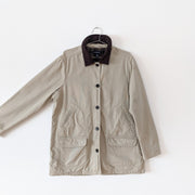 Cotton canvas field-style barn jacket, chore coat in Sand Beige (like a greige tan) with the arm extended to show sleeve and straight fit. It has dark brown buttons and dark brown corduroy lined collar. The inside of the jacket's torso is lined with green plaid flannel. This jacket has four exterior pockets: two lower patch pockets with flaps and two upper angled welt pockets. Land's End. Secondhand from Fold and Fray.