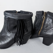 Close-up showing the leather, outer finge and interior metal zipper on Black leather heeled ankle booties with almond toe. The top of the boot is shown roll down down over western-style fringe. Sam Edelman Black Louie Boot 8M. Secondhand from Fold and Fray.