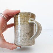 Tan/Brown Speckled Ceramic Mug, Rough Texture. Top Third has Darker Yellow Glaze. Shape Opens Wider at Mouth. Small Logo.